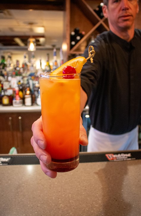 A male bartender hands someone an orange cocktail with a cherry garnish